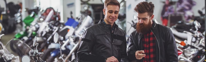 two guys in bakck leather jackets talking with about buying a motorcycle with a bunch of motorcycles around them