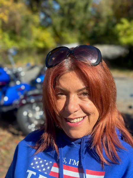 Photo of a woman wearing a sweatshirt with sunglasses on the top of her head. There's a motorcycle out of focus in the background.