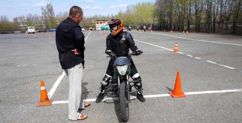 Photo of a person on a motorcycle in a parking lot with orange cones and an instructor