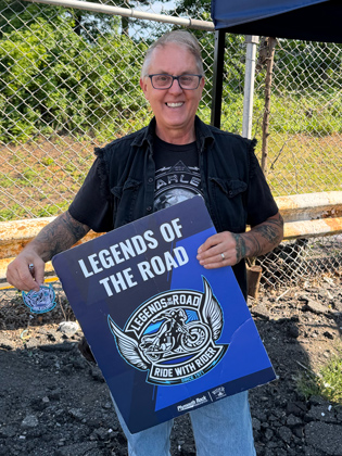 Photo of an older white man holding a sign that says "legends of the road"