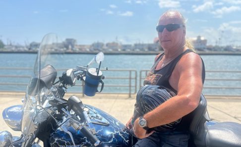 Photo of a man sitting on a motorcycle. He is parked on a path overlooking a body of water.