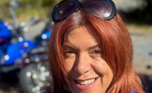 Photo of a woman wearing a sweatshirt with sunglasses on the top of her head. There's a motorcycle out of focus in the background.